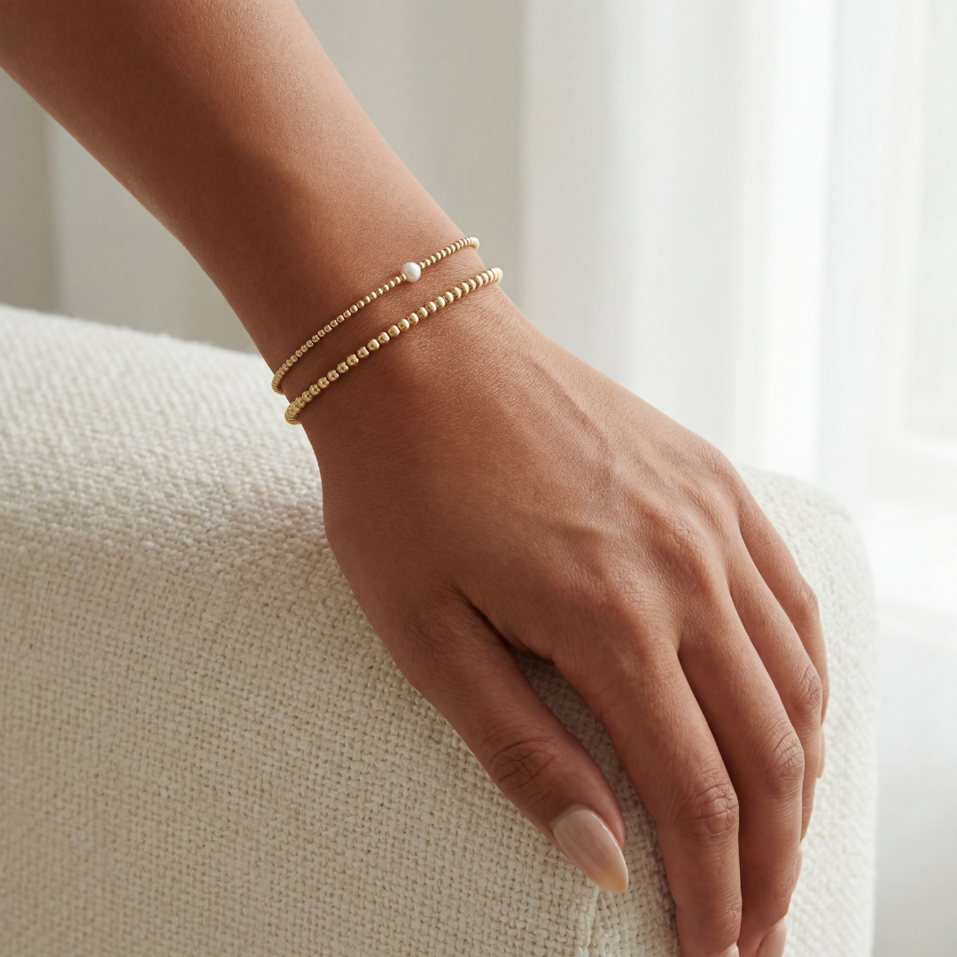 Hand wearing a gold beaded bracelet on a neutral background