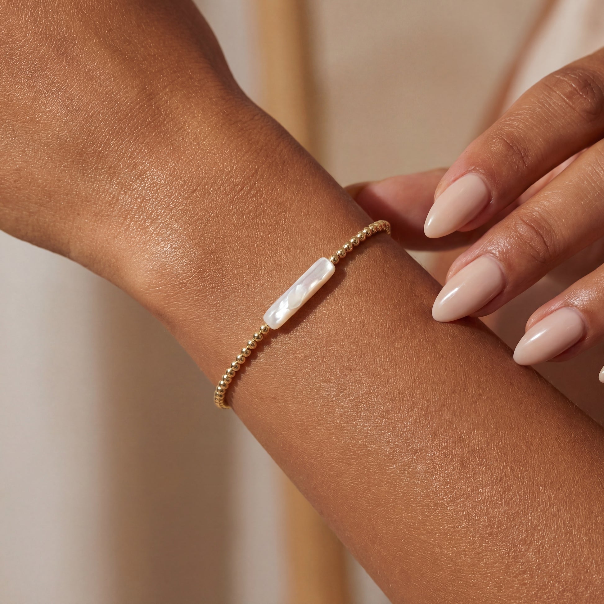 Gold beaded bracelet with a white pearl on a wrist against a beige background