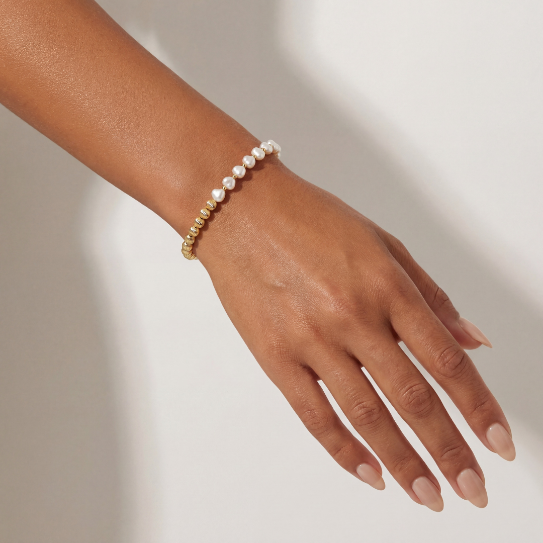 Hand wearing a pearl and gold beaded bracelet on a neutral background