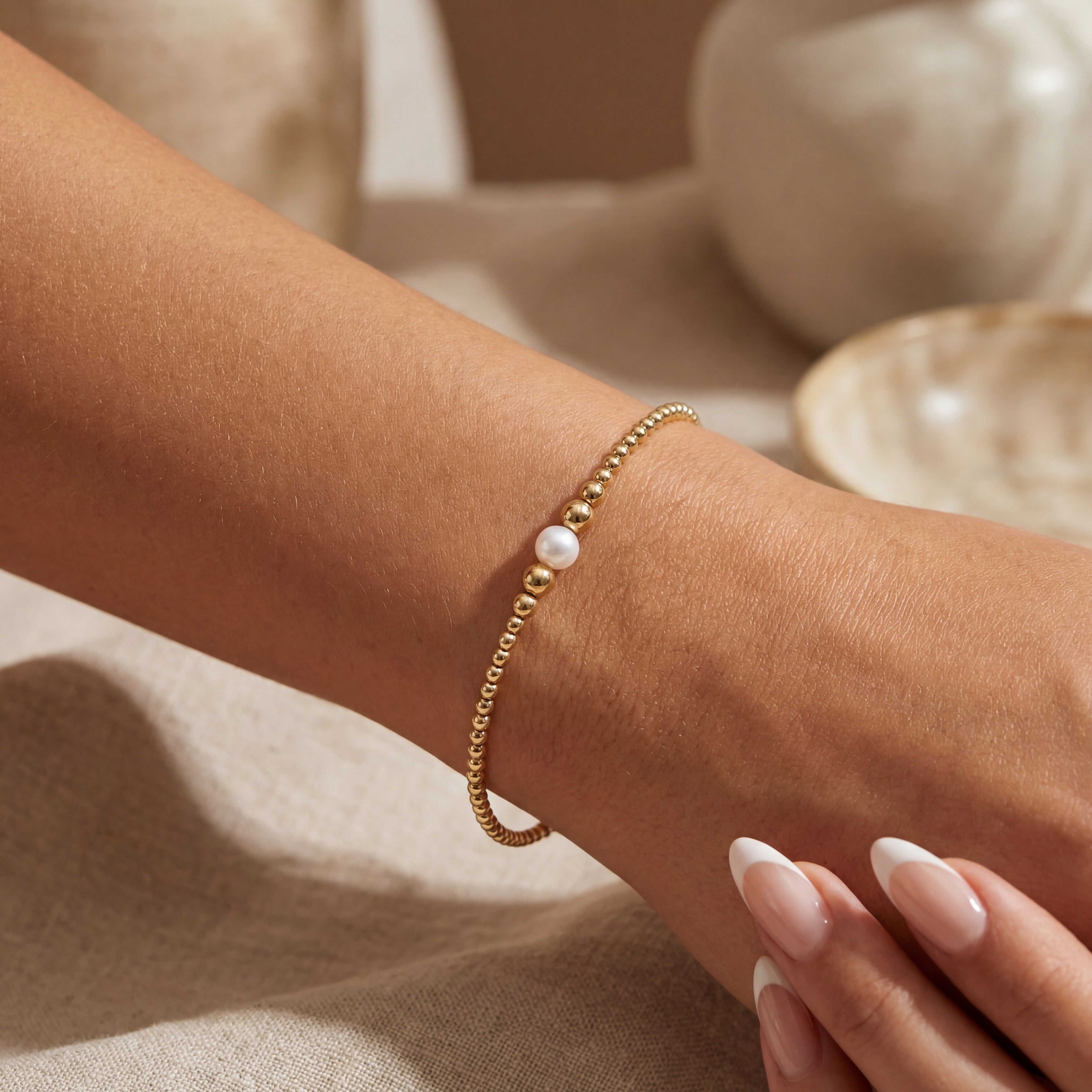 Gold beaded bracelet with a pearl on a wrist against a neutral background