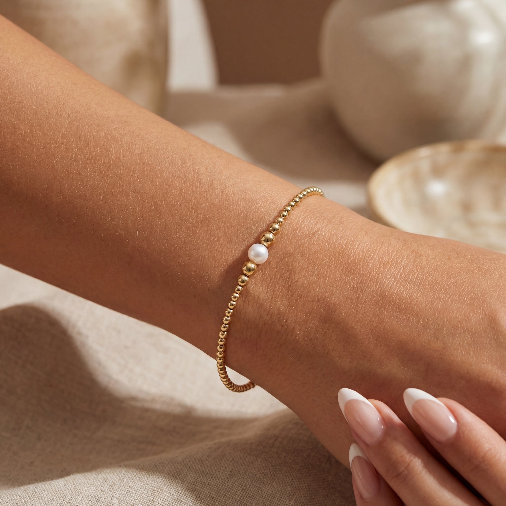 Gold beaded bracelet with a pearl on a wrist against a neutral background