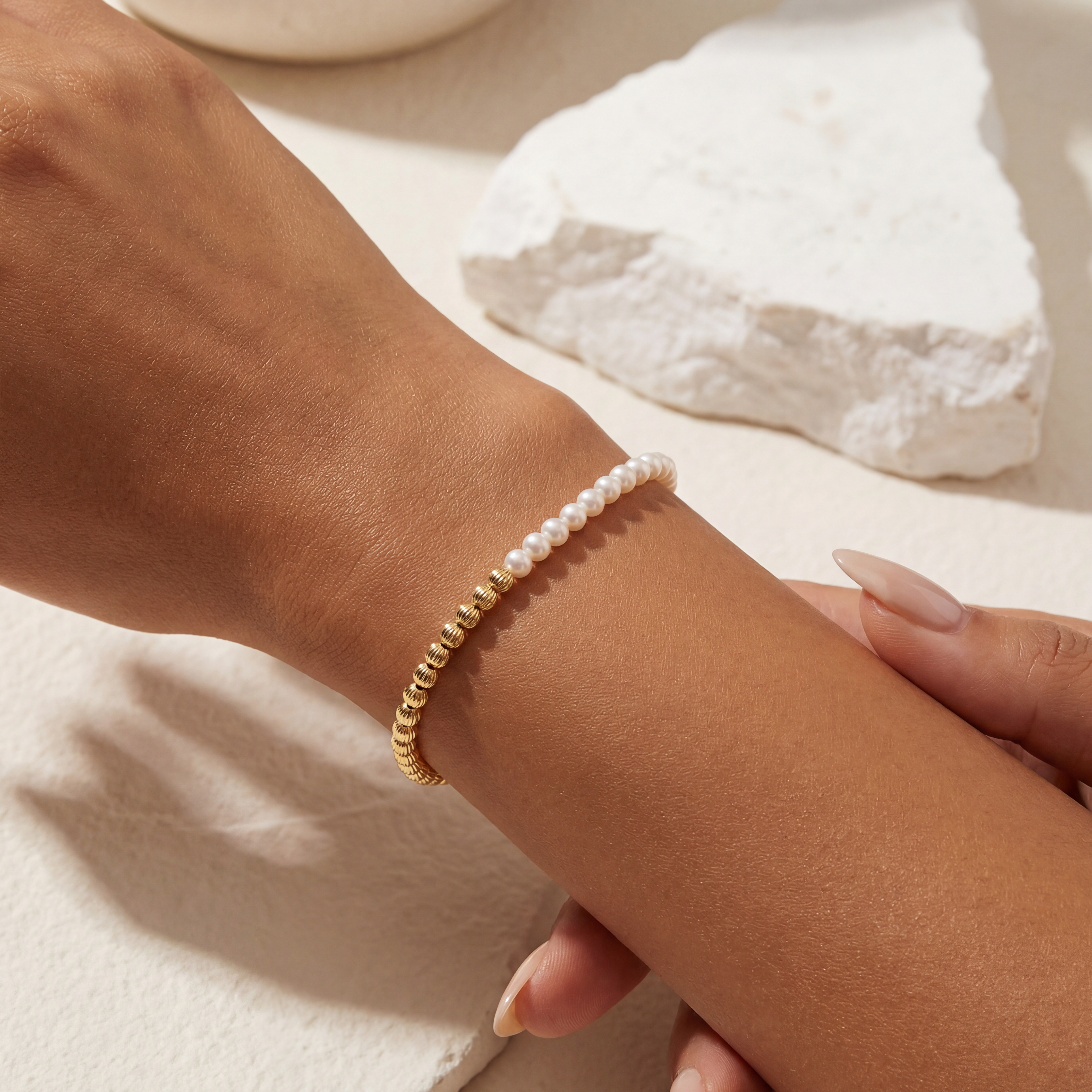 Close-up of a wrist wearing a gold beaded bracelet with a pearl on a light background