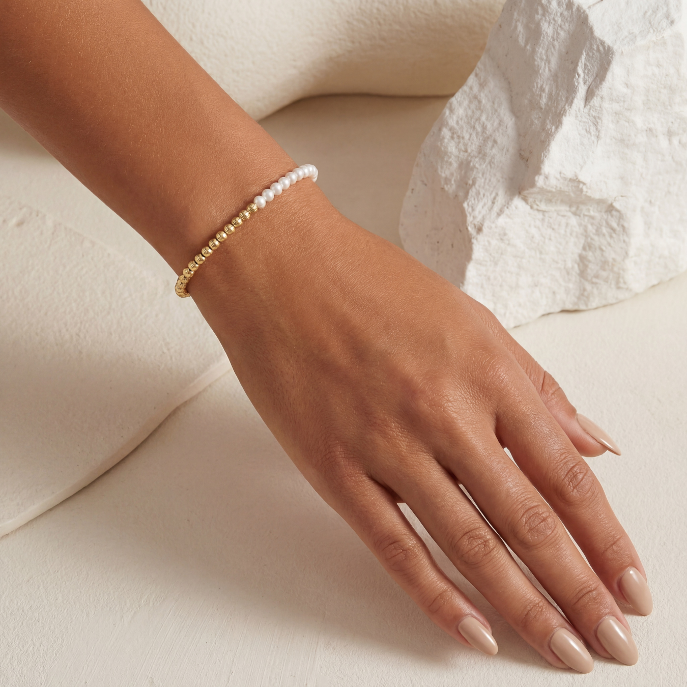 Hand wearing a gold beaded bracelet on a neutral background